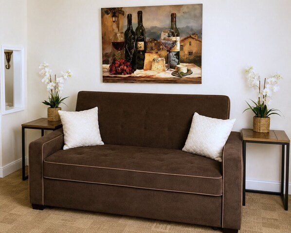 Un canapé marron agrémenté de deux coussins blancs, flanqué de deux petites tables sur lesquelles reposent des paniers en osier et des orchidées blanches. Au-dessus, on peut voir un tableau représentant des bouteilles de vin et des grappes de raisin.