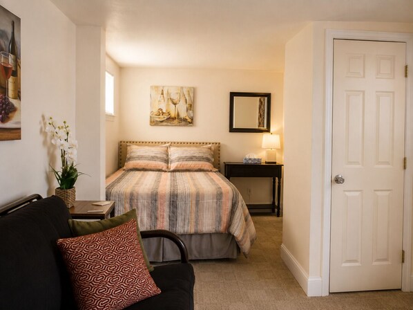A bedroom with a bed, a bedside table with a lamp, a chair, a door, and a painting on the wall.