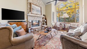 A cozy living room with a fireplace, comfortable seating, a flat-screen TV, and a large window with a view of trees. - Grand Hyatt|Creekside, multi level condo w/close access to chair 20 and views of Gore Creek (Vail)