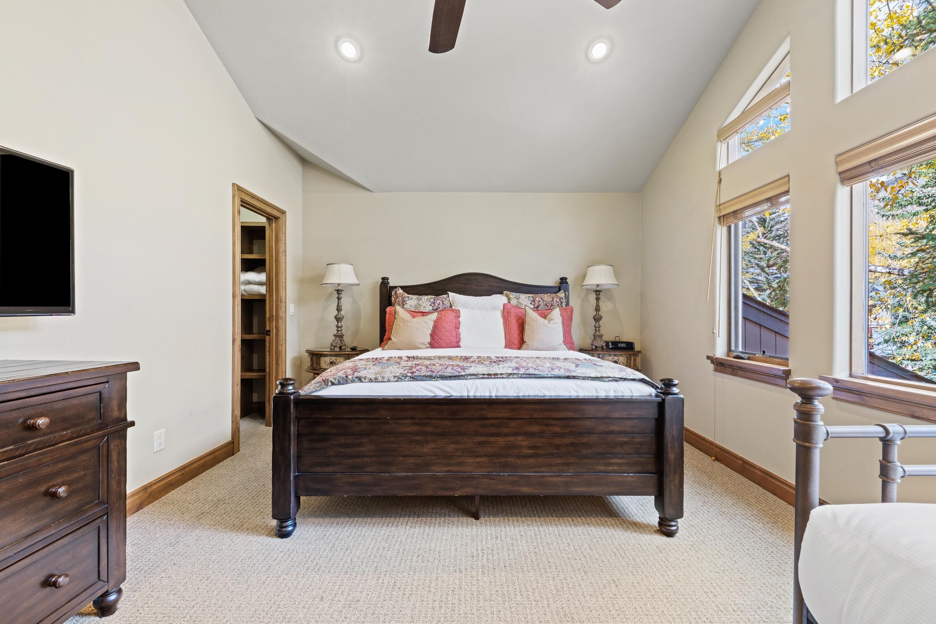 A bedroom with a dark wooden bed, two bedside lamps, a ceiling fan, a TV on the left, and a window on the right.