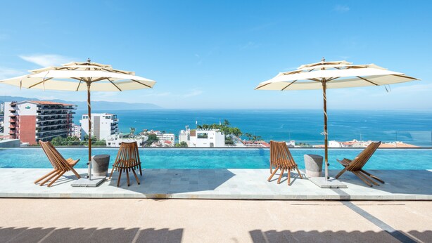 A poolside area with lounge chairs and umbrellas, overlooking a clear blue sea and a distant cityscape. - Sail View,Spacious Condo Romantic Zone Ocean Views (Puerto Vallarta)