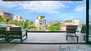 A balcony with two chairs and a table, overlooking a coastal cityscape with buildings and palm trees. - Sail View,Spacious Condo Romantic Zone Ocean Views (Puerto Vallarta)