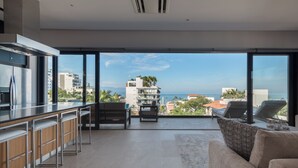 A modern kitchen with a bar area, a seating area with a sofa and chairs, and a large sliding glass door leading to a balcony with a view of the sea and buildings. - Sail View,Spacious Condo Romantic Zone Ocean Views (Puerto Vallarta)