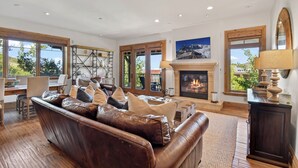 A spacious living room with a leather sofa, a fireplace, and large windows offering a view of the outdoors. - Ski-in Ski-out | Luxury | Chefs Kitchen | Garage (Park City)