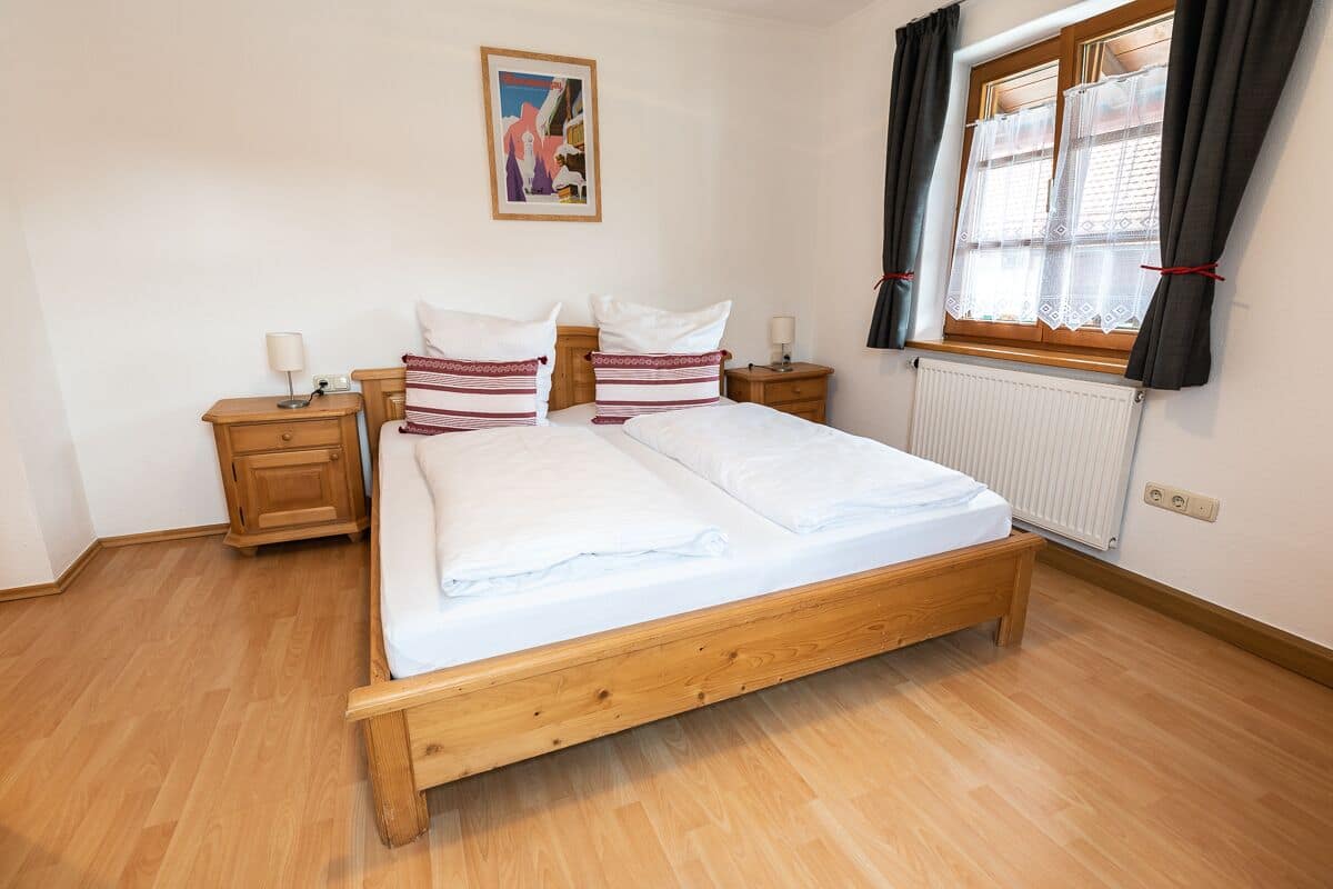 A double bed with a wooden frame, two bedside tables with lamps, a radiator, and a window with curtains.