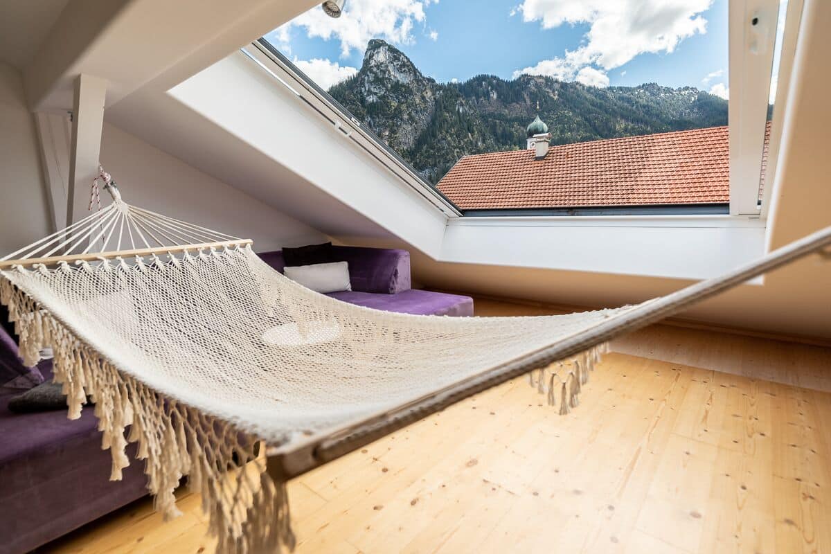A hammock in a room with a wooden floor and a view of mountains through a skylight.