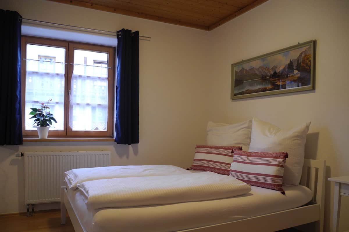 A bedroom with a bed, a window with curtains, a radiator, a plant, and a painting on the wall.