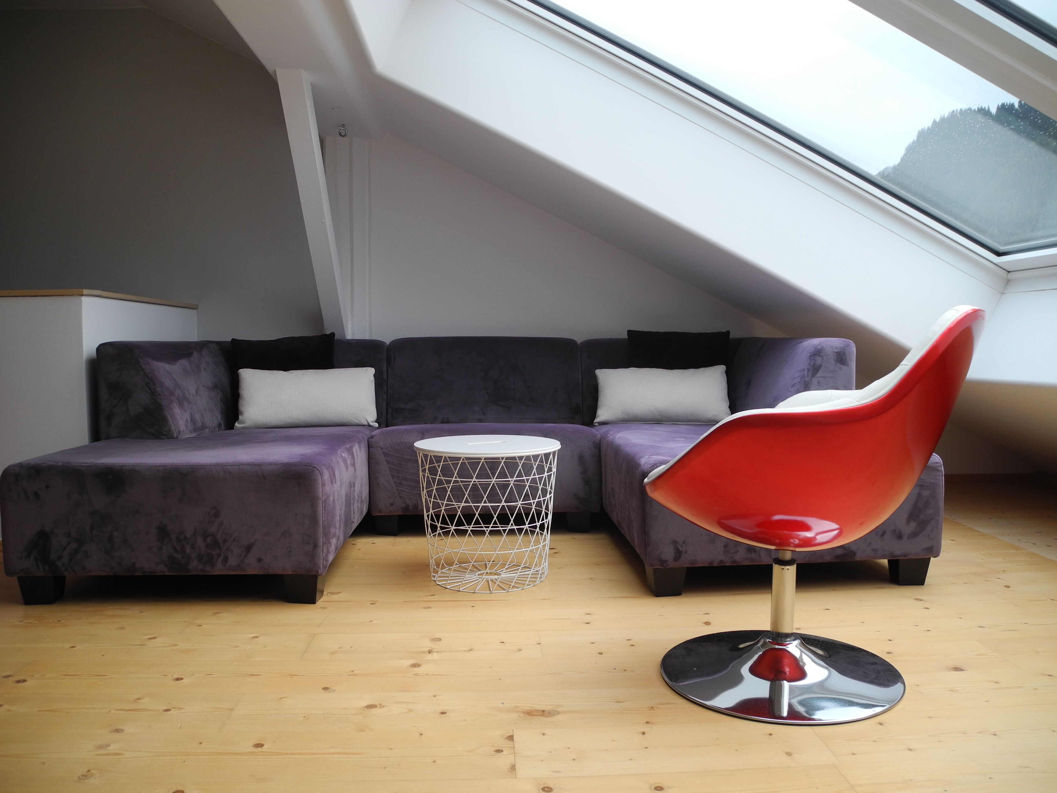 A modern loft bedroom with a sloped ceiling, a purple sectional sofa, a red chair, and a small white side table.