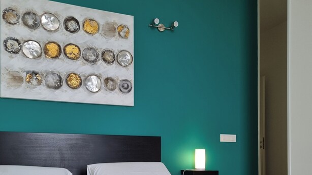 A hotel room with a teal accent wall, a bed with white and mustard pillows, a bedside table with a lamp, and a wall art piece. - Location10 (Palermo)
