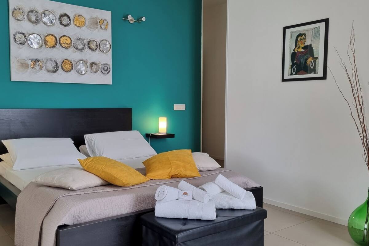 A modern bedroom with a teal wall, a large abstract painting, a framed artwork, a bed with white and yellow pillows, and a bedside table with a lamp.