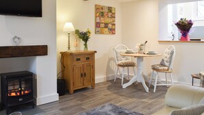 A cozy living room with a fireplace, a dining table with chairs, a TV mounted on the wall, and a window with a bouquet of flowers. - 1 bedroom accommodation in Findon, near Worthing (Findon, near Worthing)