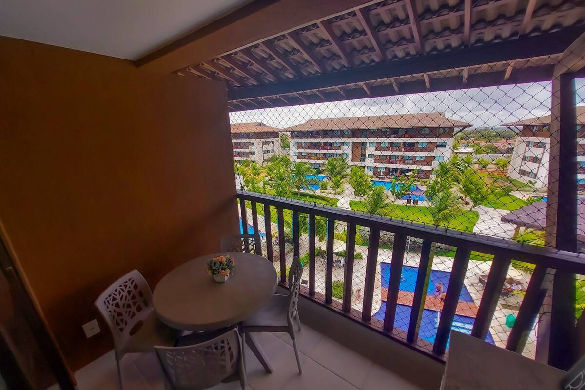 Un balcón con mesa y sillas, un pequeño jarrón de flores y vistas a una piscina y edificios.