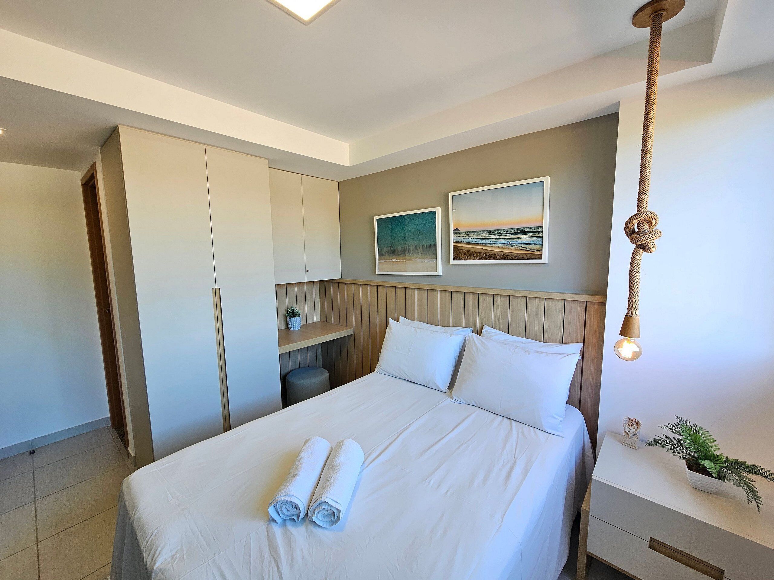A neatly made bed with rolled towels, a bedside plant, and a hanging light fixture.
