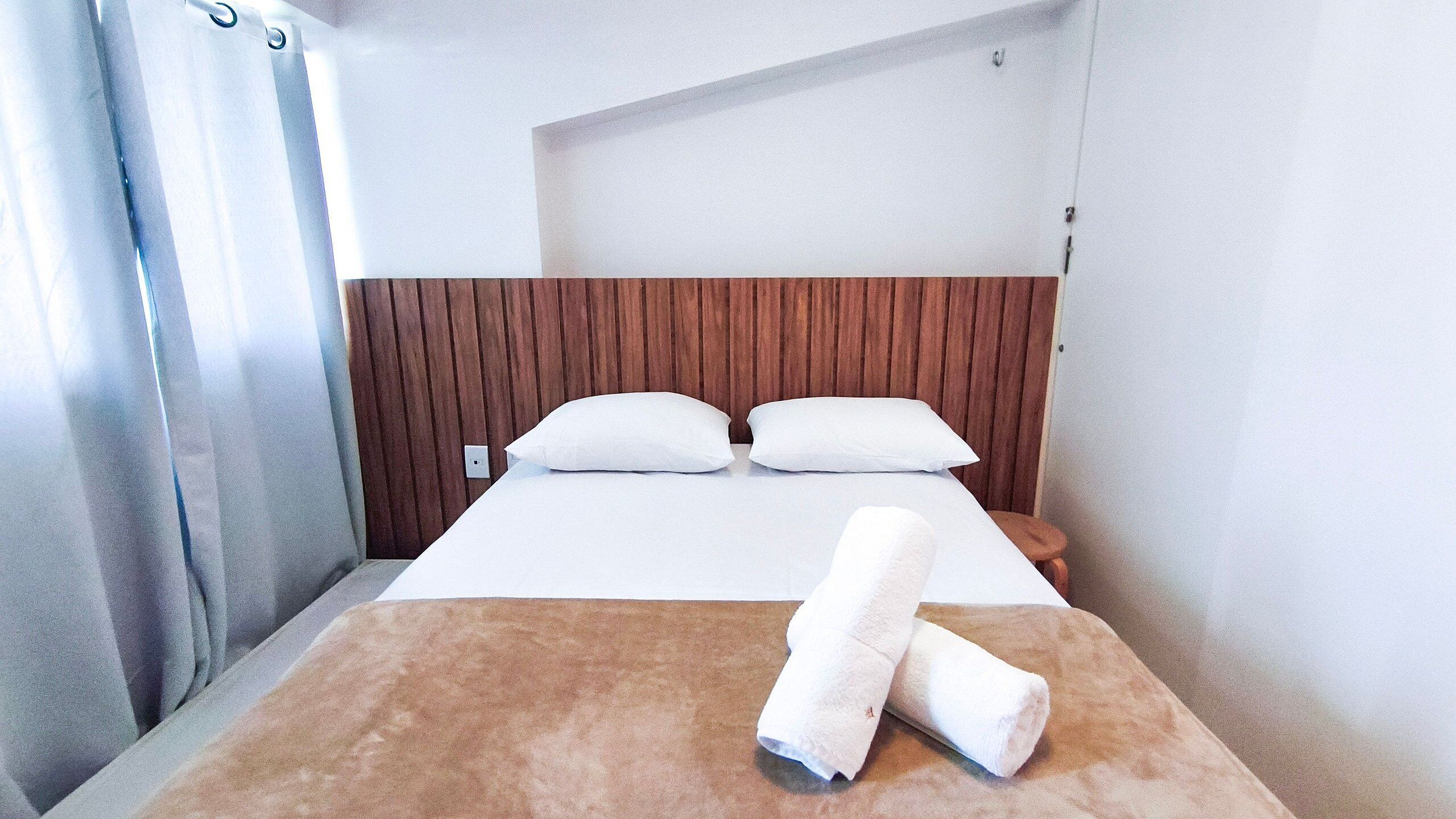 A hotel room with a wooden headboard, a bed with white linens, and a rolled towel on the bed.