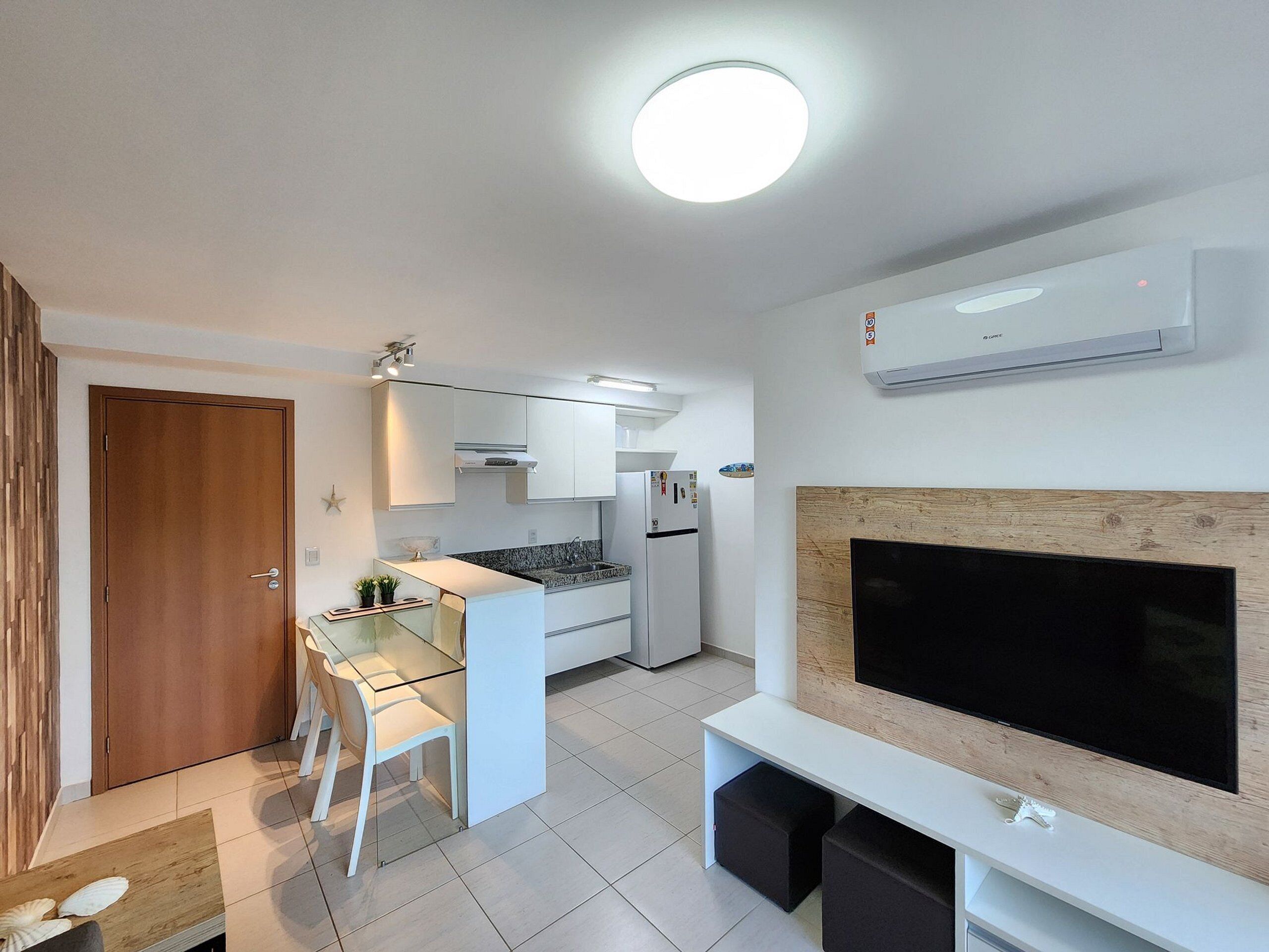 A compact living space with a kitchenette, a dining area, a flat-screen TV mounted on a wooden panel, and an air conditioning unit on the wall.