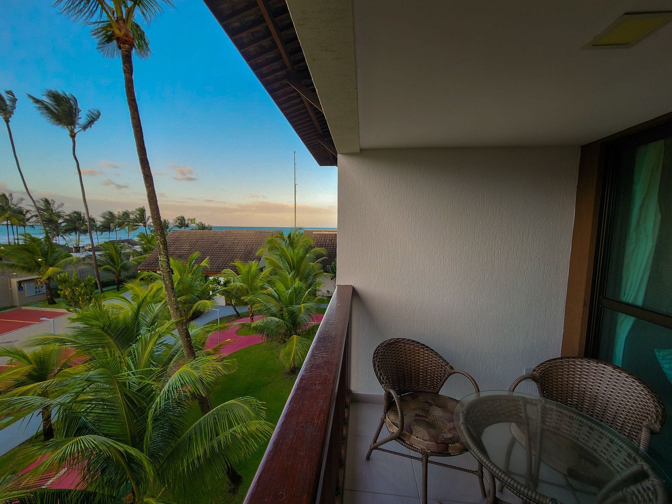 A balcony with wicker chairs, a glass table, and a view of palm trees and a beach.