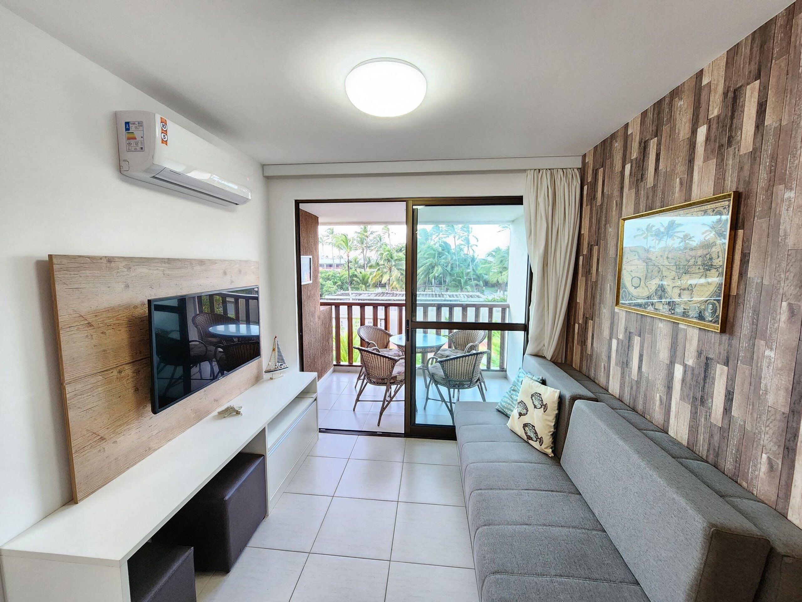 A modern living room with a flat-screen TV, a sofa, a dining area with chairs, and a balcony with a view of palm trees.