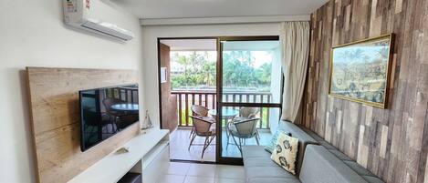 A modern living room with a flat-screen TV, a sofa, a dining area with chairs, and a balcony with a view of palm trees.