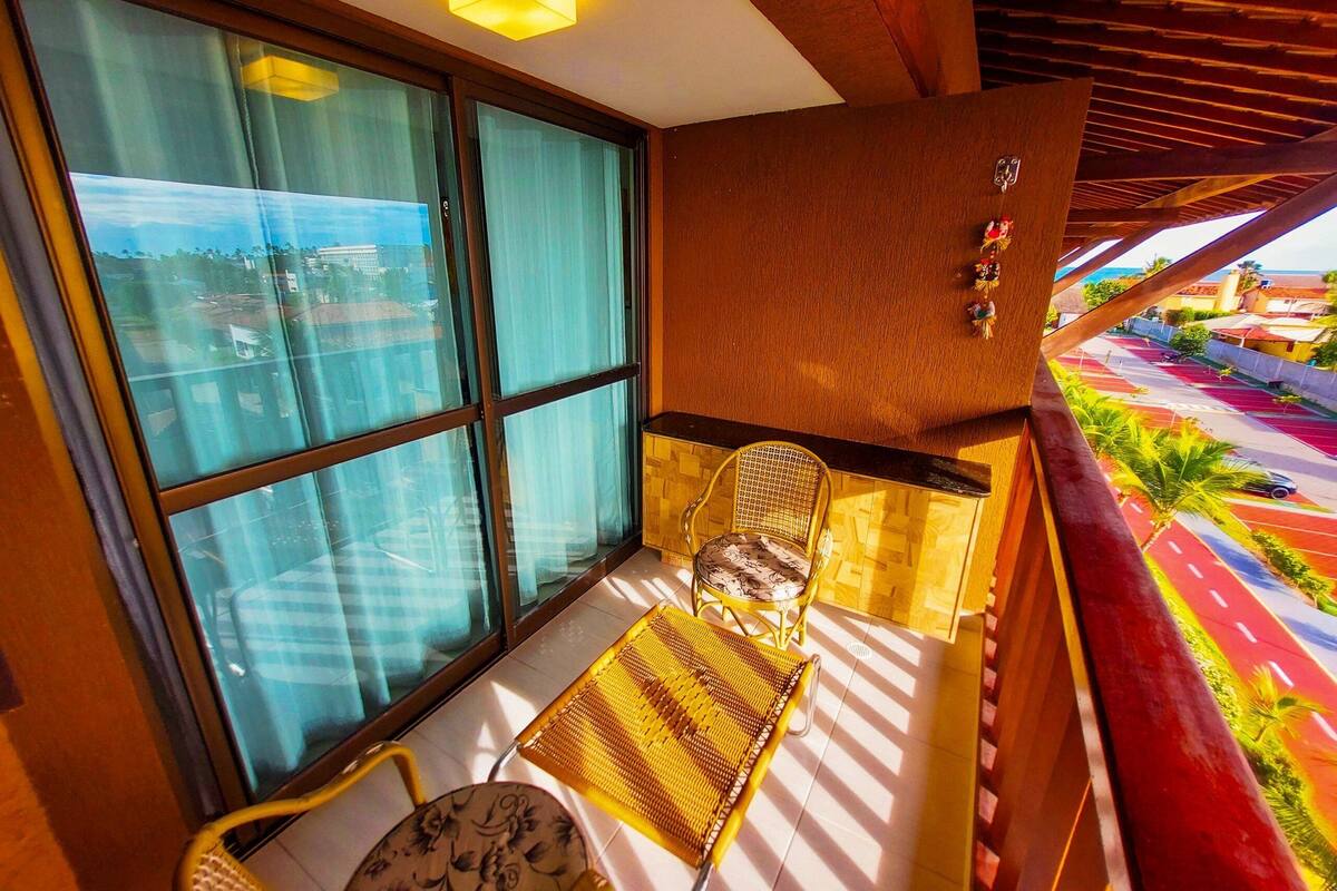 Un balcón con vista a una calle colorida, una silla, una mesita y una ventana con cortinas.