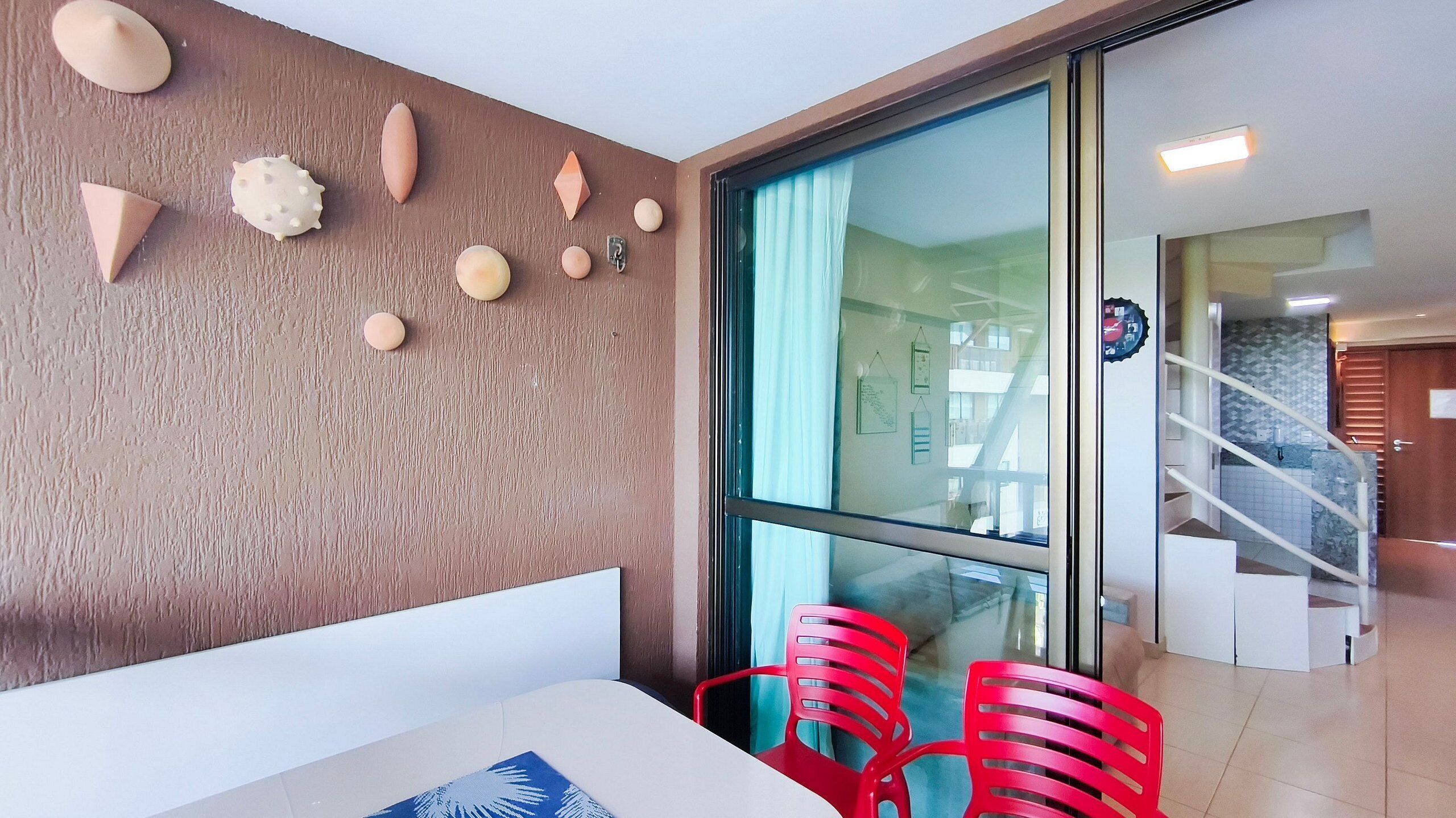Un interior moderno con zona de comedor que incluye una mesa blanca y sillas rojas, una pared con textura y objetos decorativos, y vista a una escalera que lleva a un balcón.