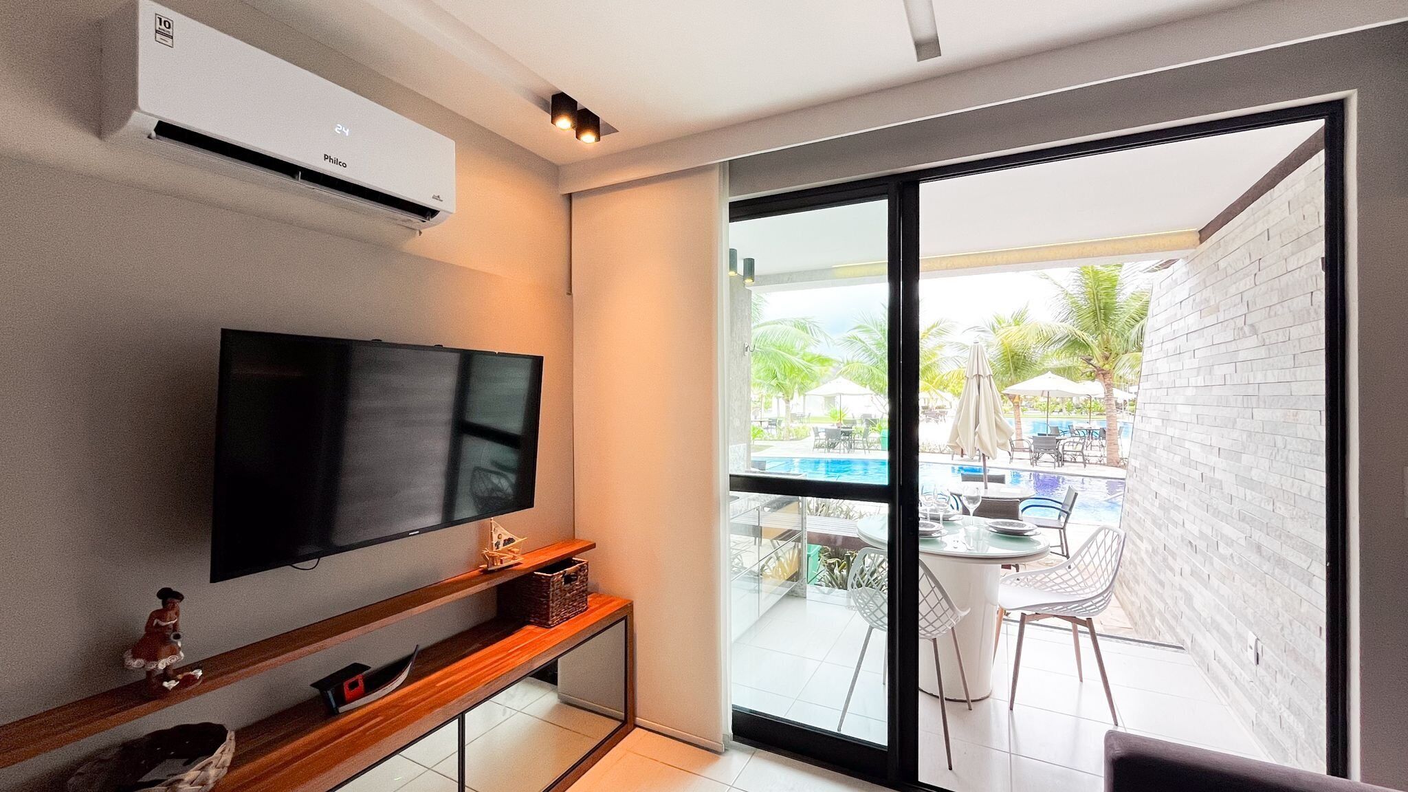 A modern living room with a flat-screen TV, a wooden console table, and a large sliding glass door leading to an outdoor pool area.
