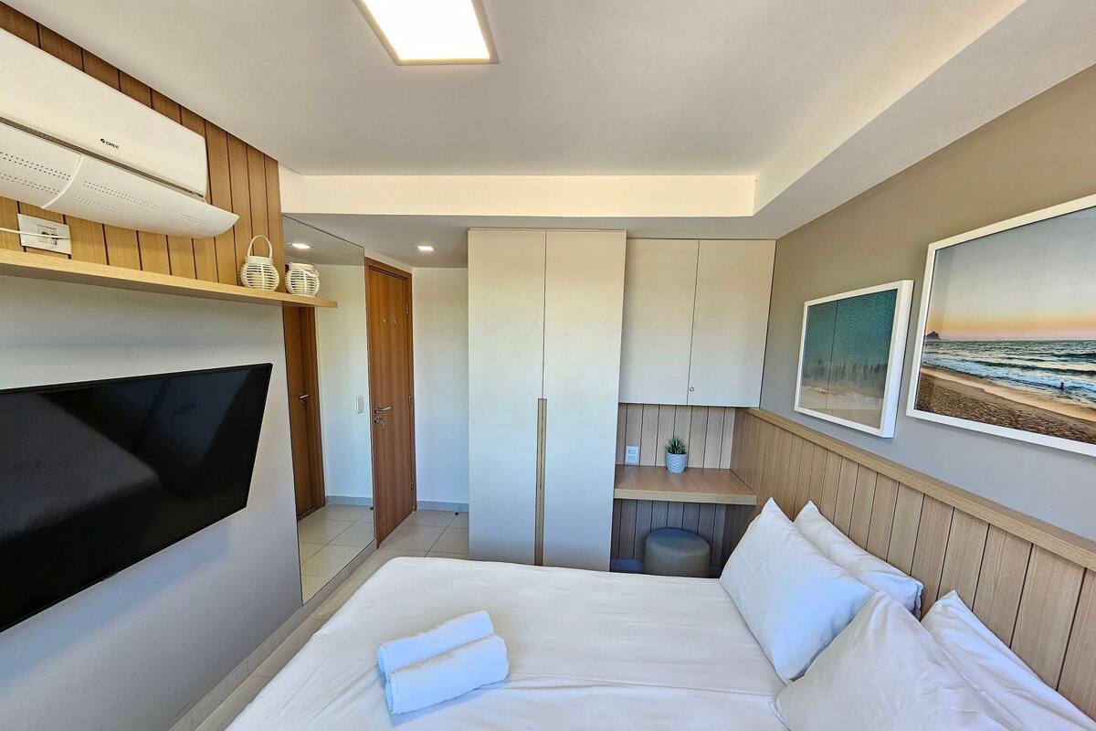 Habitación de hotel con cama, televisión, escritorio pequeño, repisa en la pared y un cuadro enmarcado de una playa.