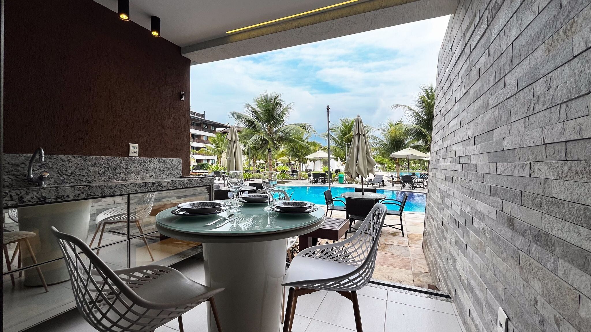 A poolside bar with a round table, chairs, and a view of a swimming pool and palm trees.