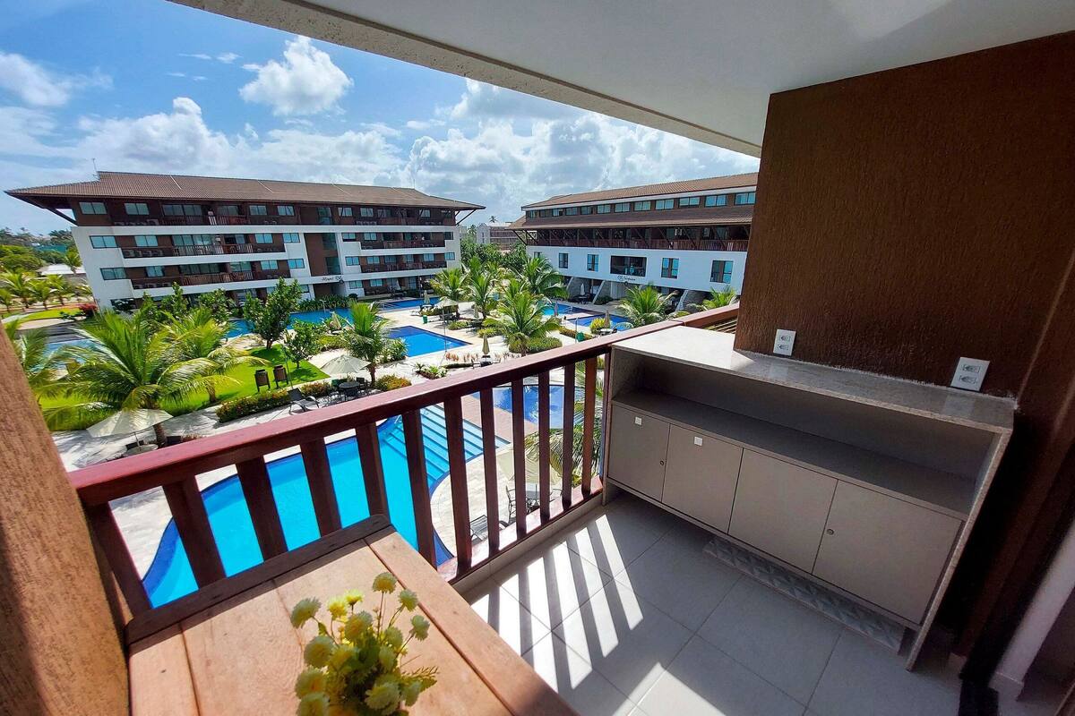 Un balcón con vistas a una piscina, un área de comedor con mesa y sillas, y un edificio con varios pisos y balcones.
