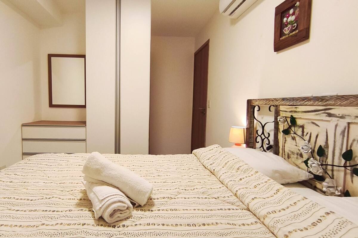 Ein Schlafzimmer mit Bett, Nachttisch, Kommode, Spiegel und einer Wandklimaanlage.