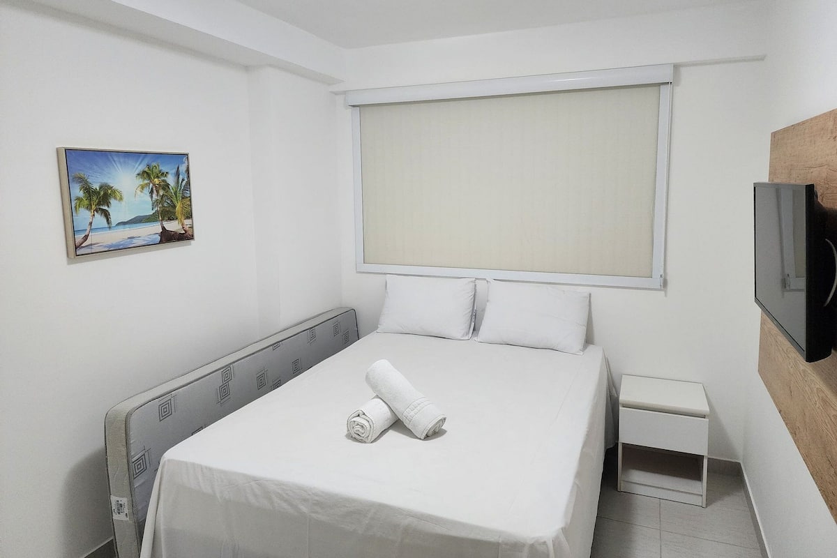 Ein Hotelzimmer mit einem Bett, einem kleinen Nachttisch, einem Fernseher und einem Bild von einer Strandlandschaft.