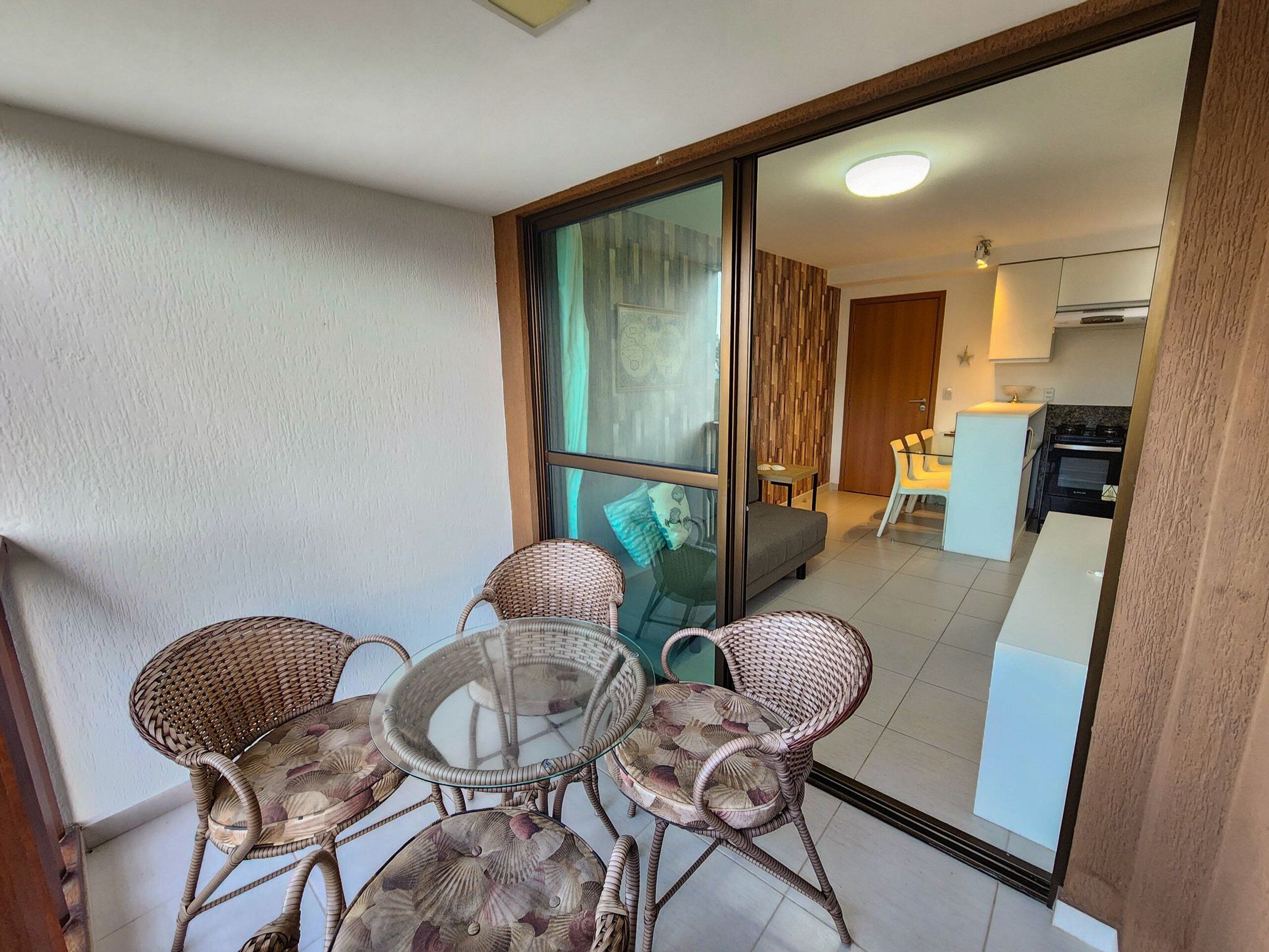 A balcony with wicker chairs and a glass table, a sliding glass door leading to a living area with a sofa and a kitchen.