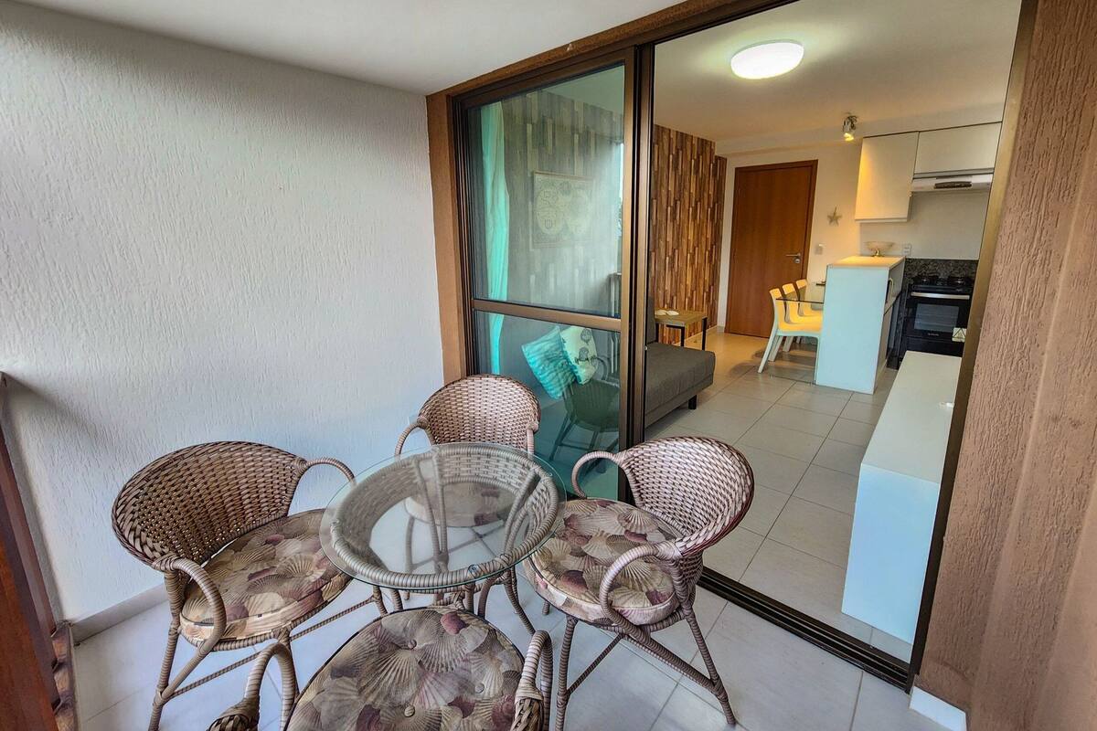 Un balcón con sillas de mimbre y una mesa de vidrio, una puerta corrediza que da a una sala de estar con sofá y cocina.