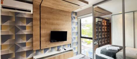 A modern hotel room with a flat-screen TV mounted on a wooden wall, a bed with a view to a pool, and a glass door leading to a balcony.