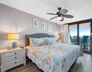 A bedroom with a bed, bedside tables, a ceiling fan, artwork on the wall, and a view through a glass door.