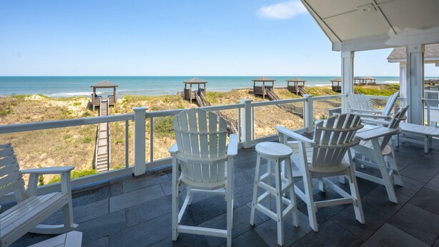 A covered outdoor area with white Adirondack chairs and a table, overlooking a beach with wooden gazebos. - Eleanor Dare 8 Bedroom Oceanfront Home, Cabana Service, Private Pool & Pet Friendly (Nags Head)