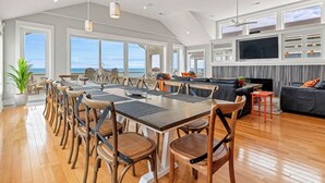 A spacious living area with a large dining table, comfortable seating, a flat-screen TV, and a view of the ocean. - Eleanor Dare 8 Bedroom Oceanfront Home, Cabana Service, Private Pool & Pet Friendly (Nags Head)
