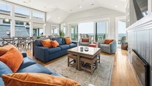 A spacious living area with a large sofa, a coffee table, and a dining table. There is a ceiling fan, large windows, and a view of the ocean. - Eleanor Dare 8 Bedroom Oceanfront Home, Cabana Service, Private Pool & Pet Friendly (Nags Head)