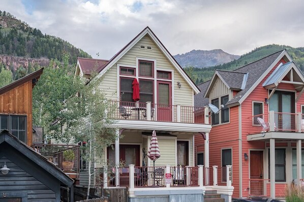 A row of colorful houses with a balcony and a porch, set against a mountainous backdrop.
