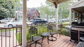 A covered outdoor patio with two metal chairs, a grill, and a view of parked cars and houses in the background. - Columbine House (Telluride)