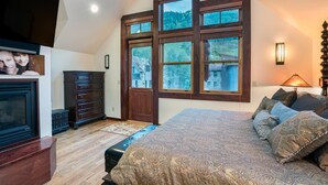 A bedroom with a large bed, a dresser, a TV, and a window with a view of a house and trees. - Columbine House (Telluride)