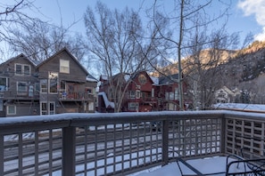 A wooden deck with a snow-covered railing, overlooking a residential area with houses and bare trees.