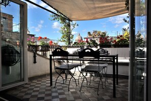 Une terrasse avec un sol à carreaux, une table entourée de chaises et une vue sur des bâtiments et des fleurs.