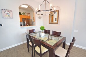 Un comedor con una mesa de vidrio, sillas de madera, un candelabro, una pequeña planta en maceta y una cocina visible a través de un arco.