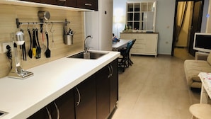 Una cocina moderna con un lavamanos de acero inoxidable, armarios de madera y una encimera con varios utensilios. Hay una zona de comedor con una mesa y sillas, y un televisor en una esquina.