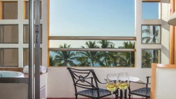 Ein Balkon mit Tisch und Stühlen, Blick auf Palmen und ein Fenster mit Meerblick.
