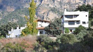 Un edificio moderno de dos pisos con techo plano y paredes blancas, rodeado de vegetación natural y un paisaje montañoso.