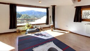 Una habitación con ventanas amplias, suelo de madera, alfombra azul y roja, y una silla blanca.