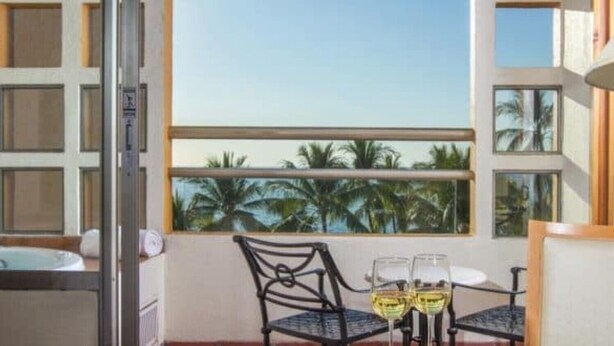 A balcony with a table and chairs, a view of palm trees, and a window with a view of the sea. - Club Regina Grand 2! (Marina Vallarta)