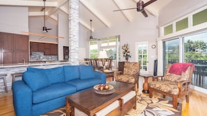 A spacious living area with a blue sofa, wooden coffee table, and patterned chairs. A kitchen is visible in the background with a stone accent wall.