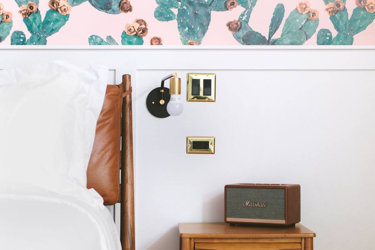 A bedroom with a bed, a nightstand with a radio, and a wall with cactus and flower wallpaper.